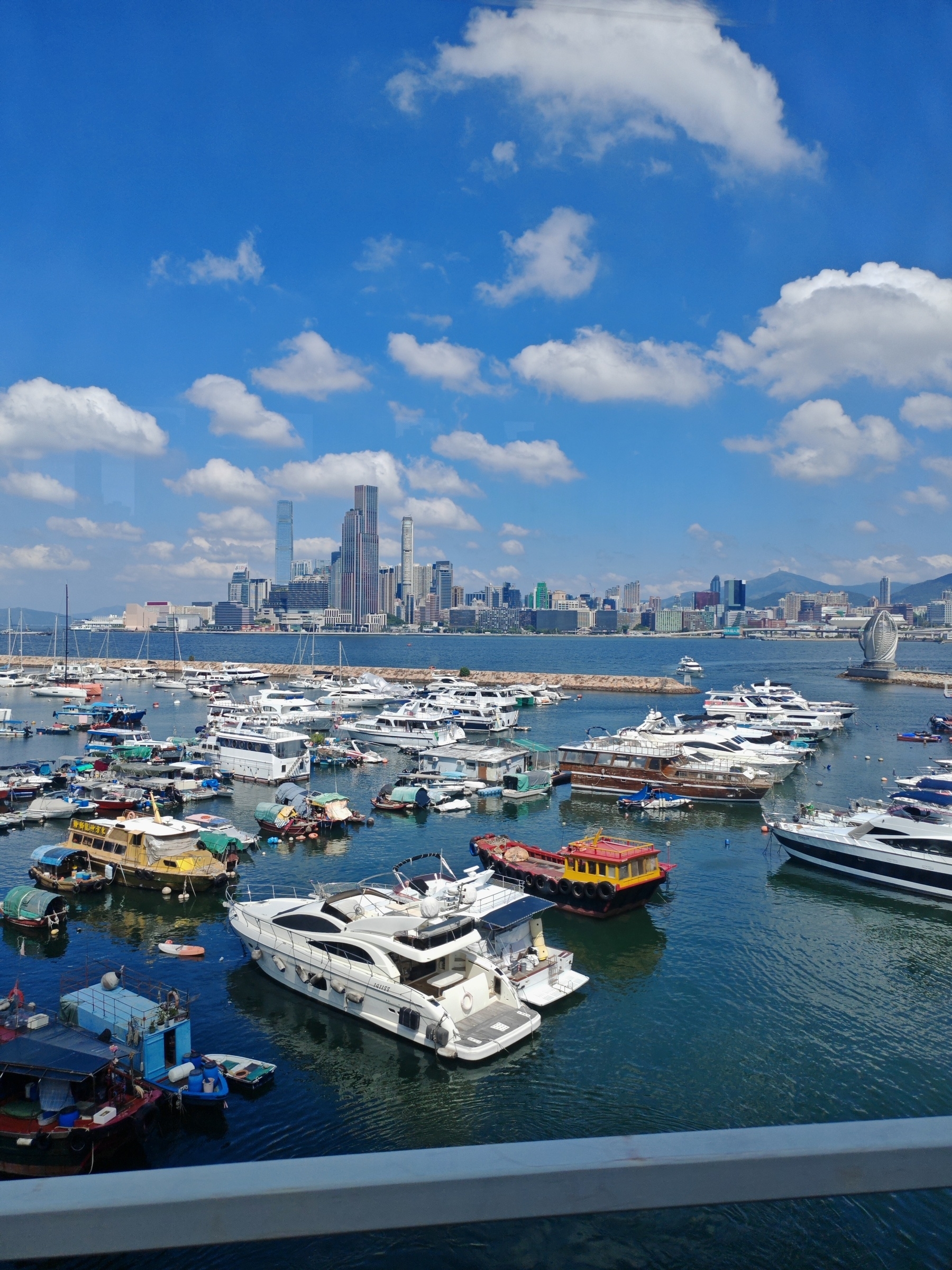 A marina filled with boats and yachts lies in the foreground, with a city skyline and blue sky with clouds in the background.